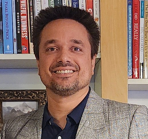 Matt Sumegi, smiling, in a grey patterned blazer over a navy shirt, in front of a bookshelf
